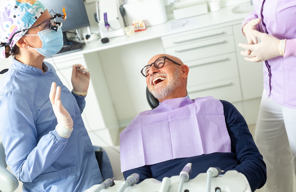 A smiling man with glasses and a lavender dental bib sits in a dental chair, laughing with two dental professionals dressed in scrubs, masks, and gloves, in a bright modern dental clinic.
