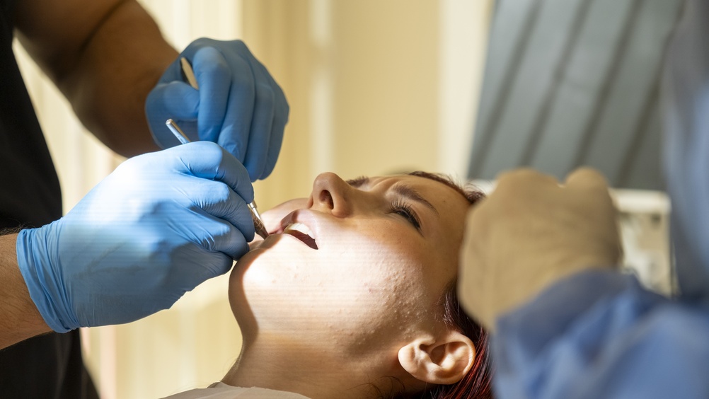 A patient lies back in a dental chair with mouth open while two gloved dental professionals use tools to examine or treat the patient's teeth in a brightly lit clinic.