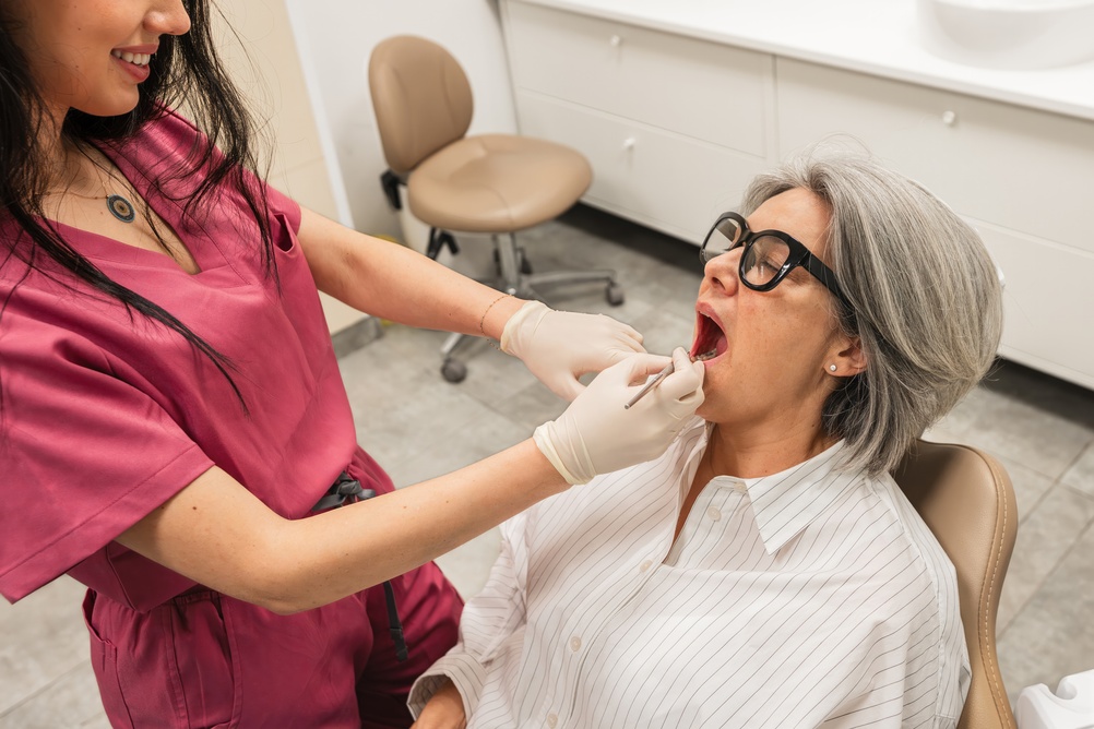 A dentist in pink scrubs and wearing gloves examines the open mouth of an older woman with gray hair and glasses, who is seated in a dental chair in a bright dental office.